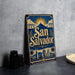 A vintage-style metal sign with the text "San Salvador" prominently displayed, featuring a sun and palm trees, is placed on a gray surface next to some black camera lenses.