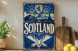 A vintage-style metal sign with the word "SCOTLAND" prominently displayed, featuring a castle, mountains, and a lion emblem. The sign is placed on a wooden surface, with a potted plant, a cutting board, and some lemons nearby.