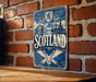 A vintage metal sign with the word "SCOTLAND" prominently displayed, featuring a shield and a deer head, mounted on a brick wall.