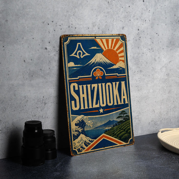 A vintage-style sign for Shizuka, a Japanese city, featuring a mountain, sun, and waves, is displayed on a gray wall next to some black camera lenses.