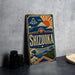 A vintage-style sign for Shizuka, a Japanese city, featuring a mountain, sun, and waves, is displayed on a gray wall next to some black camera lenses.