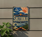 A vintage-style sign for Shizuka, a Japanese beverage, is displayed on a wall next to a plant.