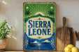 A vintage-style sign for Sierra Leone, featuring palm trees, mountains, and a sun-like design, is displayed on a shelf next to a wooden cutting board and some lemons.