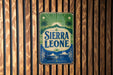 A vintage-style sign for Sierra Leone, featuring palm trees and a sunburst design, is displayed on a wooden wall.
