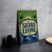 A vintage-style sign for Sierra Leone, featuring palm trees, mountains, and a sunburst design, is displayed on a gray countertop next to a stack of black camera lenses.