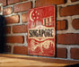 A vintage-style metal sign with the word "SINGAPORE" prominently displayed, featuring a cityscape and a crescent moon.