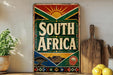 a colorful sign that says "SOUTH AFRICA" with a sun and a flower design, placed on a shelf next to a wooden cutting board and some lemons.