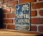 A vintage-style metal sign with the text "SOUTH CAROLINA" displayed on a brick wall.
