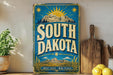 A vintage-style sign for South Dakota, featuring a mountain landscape and sun-like symbols, is displayed on a shelf next to a wooden cutting board and some kitchen utensils.