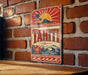A vintage-style metal sign with the word " Tahiti" prominently displayed, featuring a tropical landscape with palm trees, a sun, and a mountain in the background.