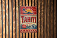 A vintage-style sign for Tahiti, featuring a tropical landscape with palm trees, a sun, and a mountain.