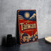 A vintage-style metal sign with the word "Taiwan" prominently displayed, featuring a sun, mountains, and a temple. The sign is placed on a gray surface, with some black camera lenses visible in the background.