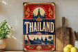 A vintage-style sign with the word "Thailand" prominently displayed, featuring a temple and other traditional Thai imagery. The sign is placed on a shelf next to a wooden cutting board and a potted plant.