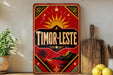 A vintage-style sign with a red background and gold accents, featuring the word "TIMOR-LESTE" prominently displayed.