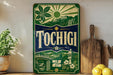 A vintage-style sign for "Tochigi" is displayed on a shelf, surrounded by various kitchen items such as a wooden cutting board, a potted plant, and a bowl of fruit.