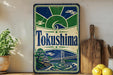 A vintage-style metal sign with the word "Tokushima" prominently displayed, featuring a bridge and a landscape scene. The sign is placed on a wooden surface, with a potted plant and a wooden cutting board nearby.