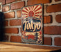 A vintage metal sign with the word "Tokyo" prominently displayed, featuring a sun and Japanese-style buildings, is mounted on a brick wall.