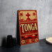 A vintage-style sign with the word "Tonga" prominently displayed, featuring a red background with palm trees and a cross, along with a sunset and island in the background.