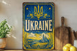a vintage-style metal sign with the word "UKRAINE" prominently displayed, along with a decorative border and a landscape illustration. The sign is placed on a wooden surface, with a potted plant and a wooden cutting board visible in the background.