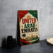 A vintage-style sign that reads "UNITED ARAB EMMIRATES" with a graphic of a city skyline and palm trees, placed on a gray countertop next to some black camera lenses.