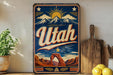 A vintage-style metal sign with the word "Utah" prominently displayed, featuring a desert landscape with mountains, a sun, and a star. The sign is mounted on a wall, and there are some kitchen utensils and a potted plant visible in the foreground.
