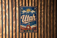 A vintage-style metal sign with the word "Utah" prominently displayed, featuring a mountain landscape and sun in the background.
