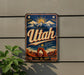 A vintage-style metal sign with the word "Utah" prominently displayed, featuring a mountain landscape and sun in the background.