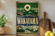 A vintage-style metal sign with the text "Wakayama" prominently displayed, featuring a scenic landscape with a temple, mountains, and a body of water. The sign is placed on a wooden surface, with a potted plant, a wooden cutting board, and some lemons nearby.