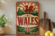 A vintage-style metal sign with a red dragon and the word "Wales" prominently displayed, placed on a wooden surface next to a wooden cutting board and some lemons.