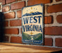 A vintage-style metal sign displaying the text "WEST VIRGINIA" with a mountain landscape and a bridge, mounted on a brick wall.