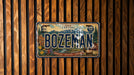 A vintage Montana license plate with the word "BOZEMAN" prominently displayed, featuring a scenic mountain landscape and various Montana-themed elements.
