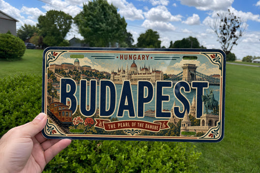 A person is holding a license plate with the word "BUDAPEST" prominently displayed, featuring an illustration of the city of Budapest.
