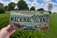 A person is holding a Michigan license plate that says "Mackinac Island" with a vintage illustration of Mackinac Island and its surrounding area.