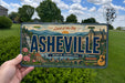 A person is holding a license plate with the text "Land of the Sky" and "Ahesville North Carolina" on it, with a scenic background of mountains, buildings, and a blue sky.