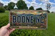 A person is holding a license plate with the text "BOONE NC" on it, which is set against a scenic mountain landscape.