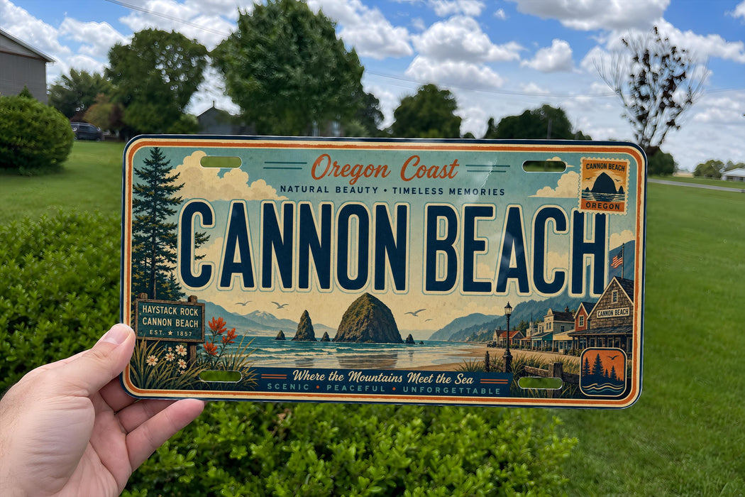 A person is holding a license plate that says "Oregon Coast Cannon Beach" with a scenic mountain landscape and ocean view on it.