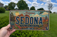 A person is holding a license plate with the word "SEDONA" prominently displayed, featuring a desert landscape with mountains, cacti, and a clear blue sky.