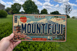 A person is holding a framed license plate with the word "MOUNT FUJI" on it, which is a Japanese license plate. The plate features a scenic image of Mount Fuji and other Japanese landmarks.
