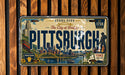 A vintage license plate from Pittsburgh, Pennsylvania, with a cityscape and landmarks depicted on it.