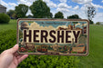 A vintage license plate featuring the word "HERSHEY" and various landmarks associated with the brand, such as a train, roller coaster, and other iconic structures.