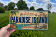 A vintage license plate from Paradise Island, featuring a tropical beach scene and palm trees.