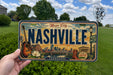 A person is holding a license plate from Nashville, Tennessee, which features a vintage design with a guitar, flowers, and a cityscape.