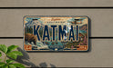 A vintage license plate with the word "KATMAI" prominently displayed, featuring a scenic mountain landscape with a bear and a plane in the background.