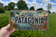 A person is holding a license plate with the word "PATAGONIA" prominently displayed on it. The license plate also features a scenic illustration of Patagonia, including mountains, a lighthouse, and other natural elements.