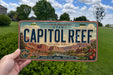 A person is holding a license plate with the text "CAPITOL REEF" and an illustration of a desert landscape.