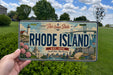 A person is holding a Rhode Island license plate with a scenic illustration of the state's landmarks and natural beauty.