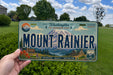 A person is holding a license plate with the text "Mount Rainier" and various scenic elements, such as a mountain, trees, and a road, against a backdrop of a grassy field and a tree.
