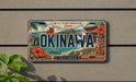 a decorative license plate with the text "OKINAWA" prominently displayed, surrounded by various Japanese-themed illustrations such as a temple, flowers, and a boat.