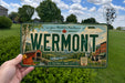 A person is holding a Vermont license plate with a scenic background featuring a barn, trees, and a river.