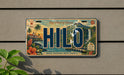 A license plate with the word "HILO" prominently displayed, featuring a tropical island landscape with palm trees, waterfalls, and other scenic elements.